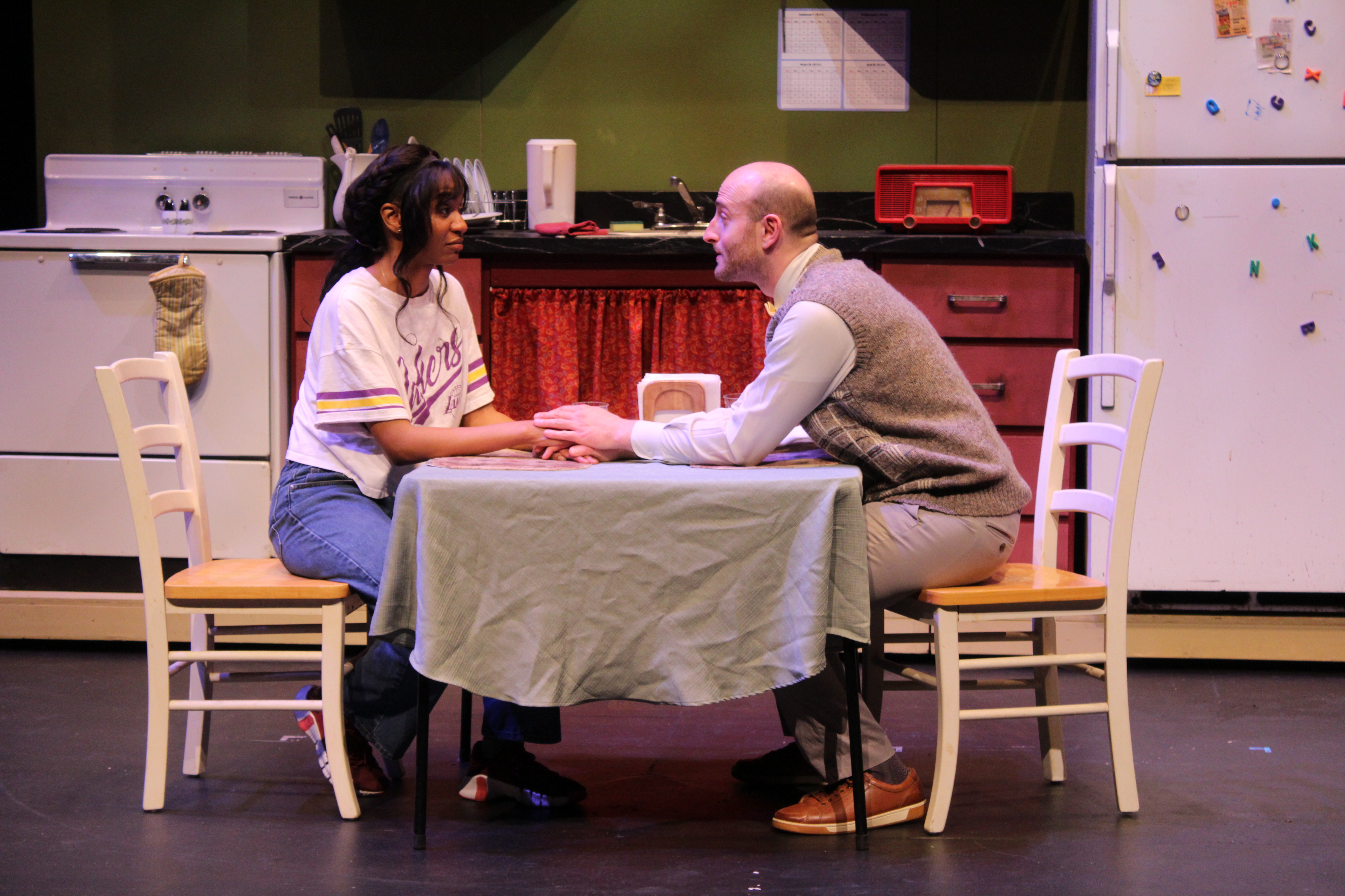 two performers sit at a table on stage while acting out in play.