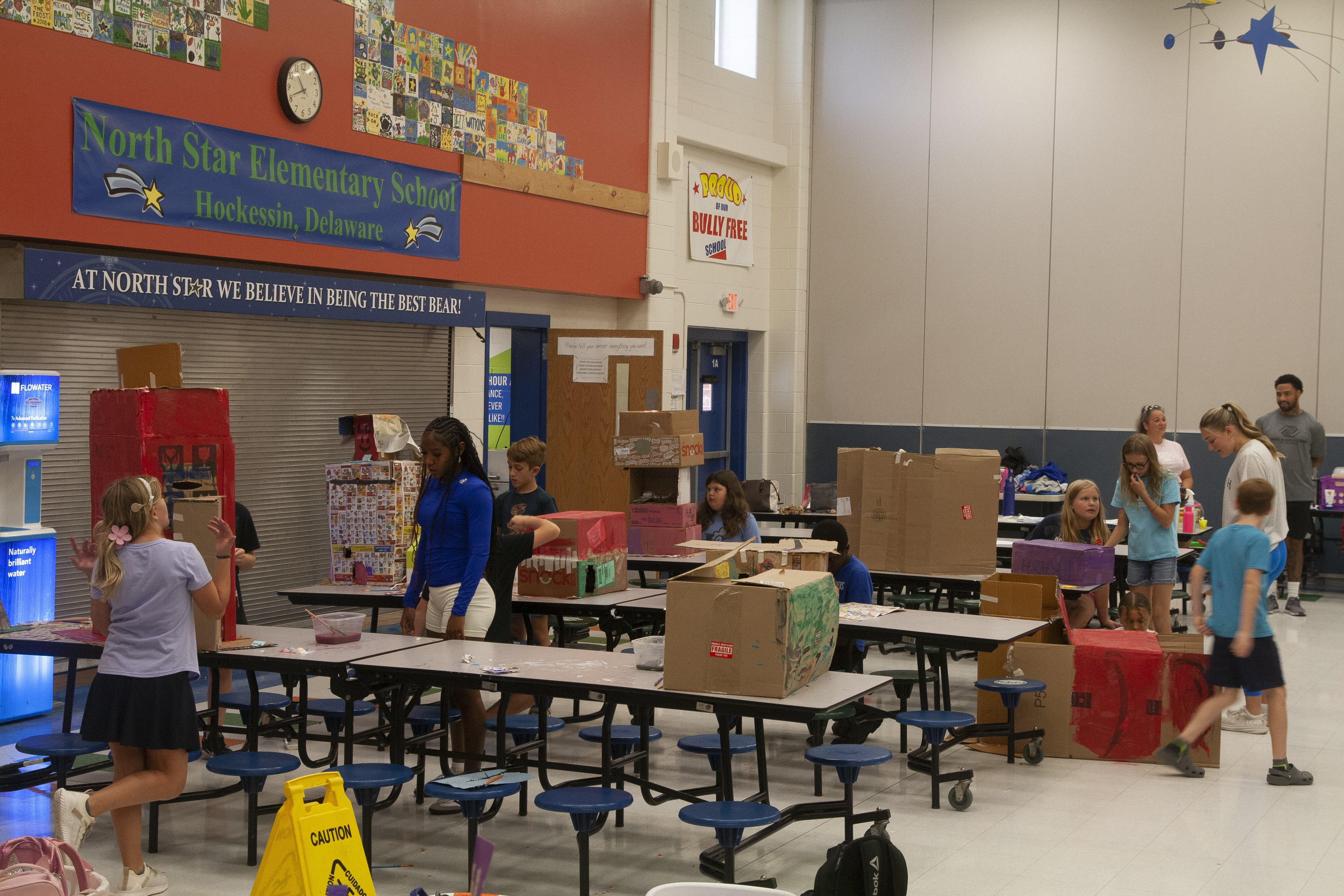 Teen Instructors look on while children pose in front of their arts and crafts in a elementary school auditorium.