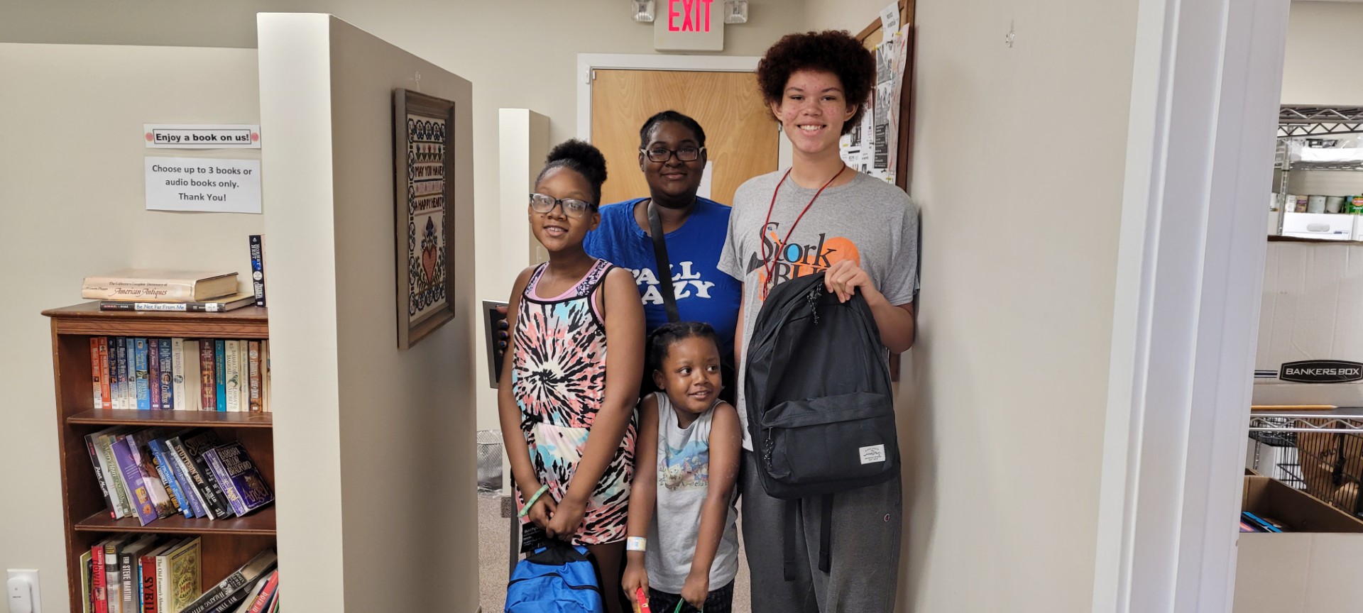 four individuals stand in a hallway holding new bookbags and recently donated items while posing for the camera.