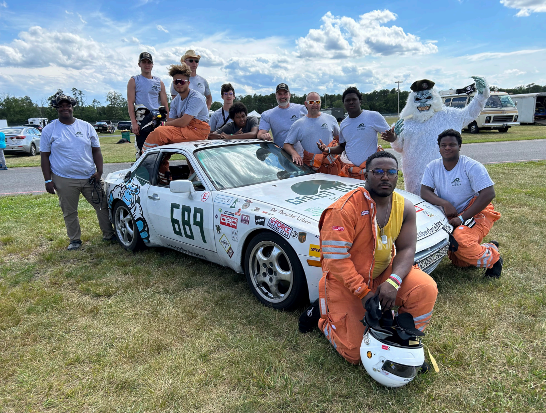 A group of teenagers stand around and huddle by a street racing car at a race track.