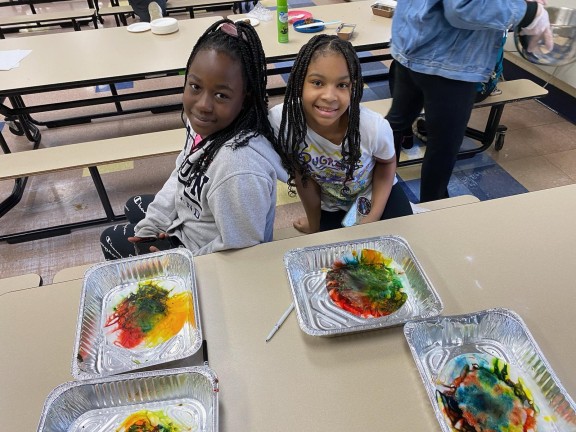 students in a cafeteria with their arts and crafts in front of them.