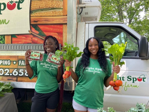 two persons holding vegetables in front of a Bright Spot farms van.