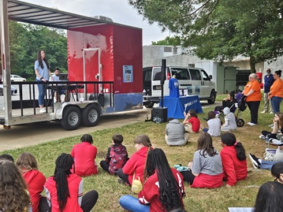 students sit while listening to a play in an outdoor open field.